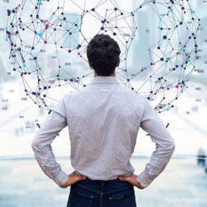 A man stands with his hands on his hips, facing a large digital network visualization with interconnected nodes and data points displayed in front of him. The image conveys a focus on data analysis, technology, or networking. Photo credit: Shutterstock