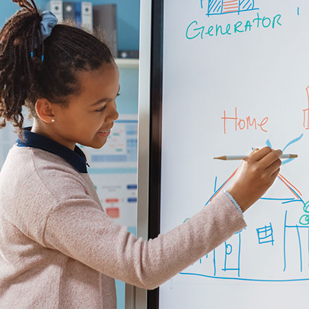 A young girl is standing at a digital whiteboard, drawing a diagram of a house and a generator. She is holding a marker and appears to be explaining her drawing, which is part of a classroom or educational setting. Credit to Shutterstock.