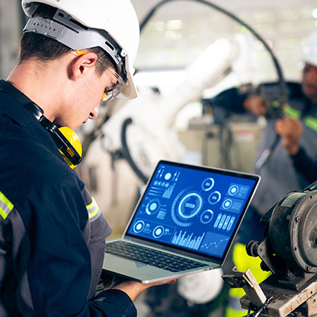 A male engineer wearing a hard hat and protective gear is using a laptop with a futuristic digital interface displayed on the screen. He appears to be inspecting machinery in an industrial setting while another worker in the background operates equipment. Credit to Shutterstock.