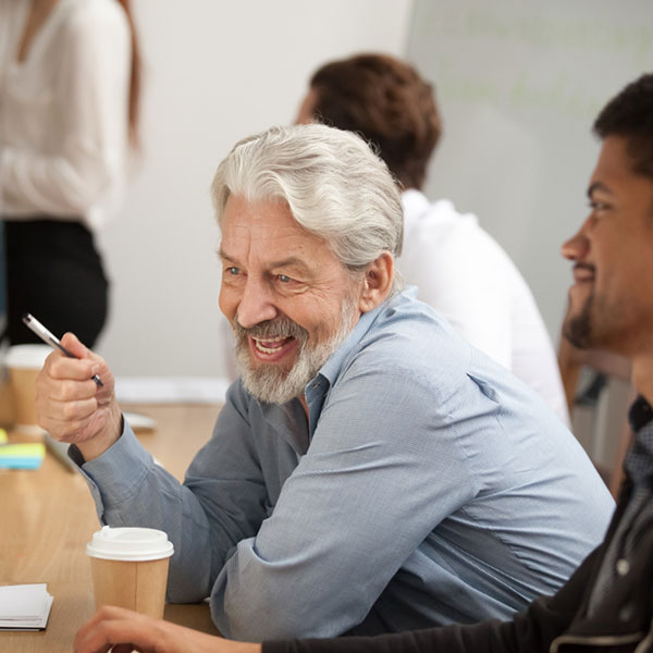 An older man with gray hair and a beard smiling while holding a pen, engaged in conversation during a meeting. A coffee cup sits on the table in front of him, and others in the room appear to be listening attentively. The atmosphere is relaxed and friendly. Photo credit: Shutterstock