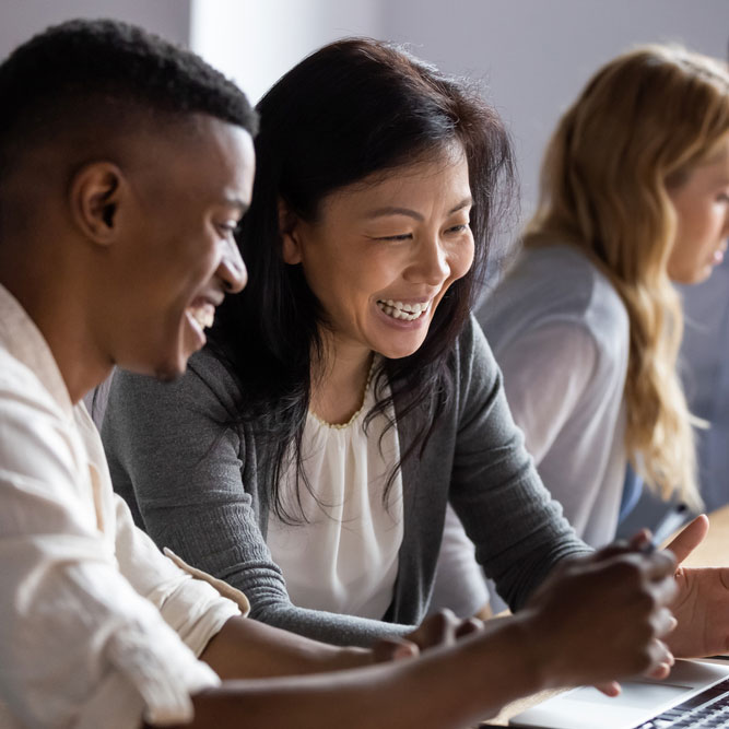 A diverse group of colleagues smiling and working together, with one person pointing at a device screen. The atmosphere appears collaborative and friendly, with a focus on teamwork. Photo credit: Shutterstock