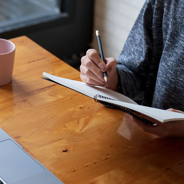 A person writes in a notebook with a pencil while seated at a wooden table. A laptop and a mug are visible on the table, suggesting a workspace or study environment. The image emphasizes focus and productivity. Photo credit: Shutterstock