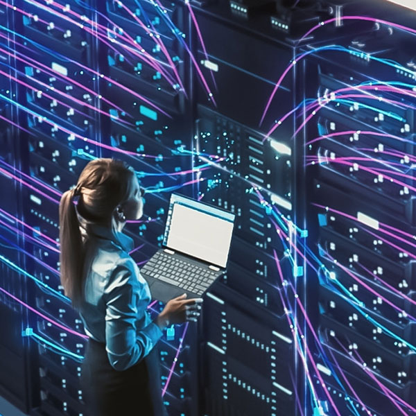 A woman stands in a data center holding a laptop, analyzing a server rack. Digital lines of data flow through the servers, symbolizing network activity and data management. The image emphasizes technology, cybersecurity, or IT infrastructure. Photo credit: Shutterstock