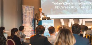 Mark Thompson stands at a podium during a conference titled “ND2024: Public Services in the Age of AI,” with an audience seated at tables. A banner displaying event sponsors is visible on the left, and a large screen behind the speaker highlights the event’s social media links using hashtags #DLWeek and #DigiLeaders. Credit: Digital Leaders