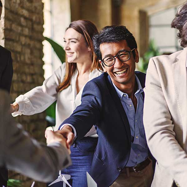 A cheerful business professional shaking hands with a colleague during a meeting. Other team members in the background are smiling, reflecting a positive and collaborative atmosphere in a modern office setting. Photo credit: Shutterstock.