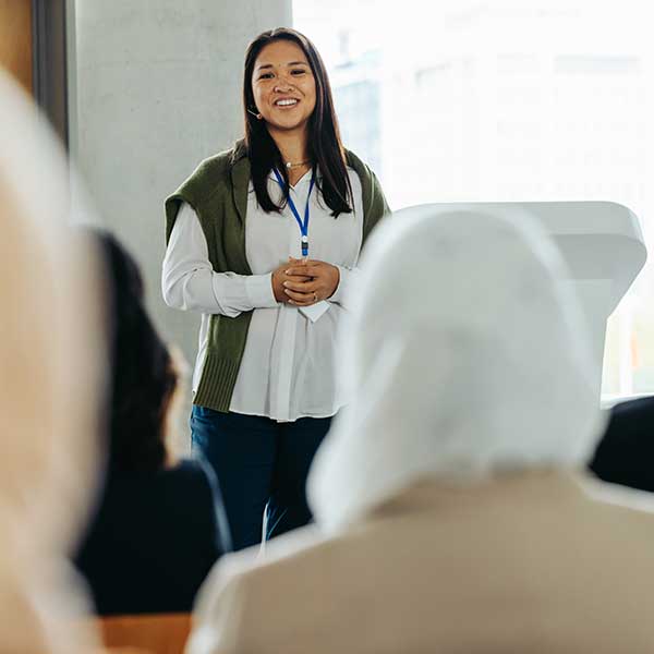 A woman is standing at a podium, smiling as she presents to an audience. The setting appears to be a conference or seminar, with people seated and listening attentively in the foreground. The environment is professional, with a large window in the background allowing natural light to enter. Photo credit: Shutterstock.