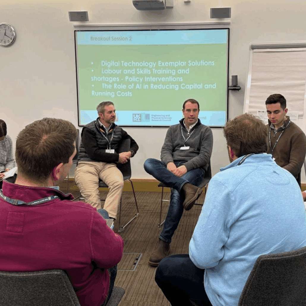 Group discussion during a breakout session on digital technology and AI, with a presentation slide listing topics such as "Digital Technology Exemplar Solutions" and "The Role of AI in Reducing Capital and Running Costs." Participants are seated in a circle, engaged in conversation.
