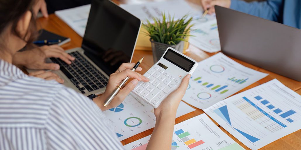 A close-up of a person's hands working at a desk. One hand is holding a pen and a white calculator, while the other is typing on a laptop keyboard. The desk is cluttered with papers displaying charts and graphs, a small potted plant, and a notebook with a pen.