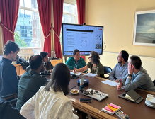 Seven people seated around a conference table in a meeting room with a large screen showing text and diagrams; red curtains cover large windows, and a framed picture hangs on the wall. Notebooks, pens, and devices are on the table.