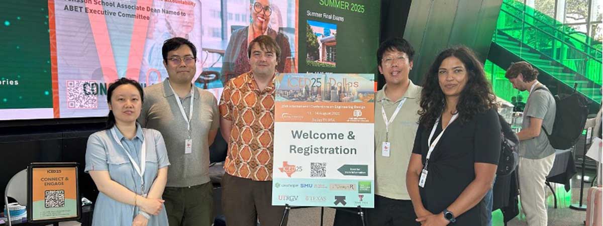 Group of five people posing indoors beside a “Welcome & Registration” sign; digital screen behind them shows info about Dr. Yvette E. Pearson and Summer 2025 Final Exam schedule