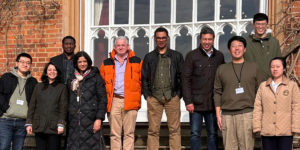 Group photo of eleven DIGIT Lab team members standing outdoors in front of a brick building and large arched window, wearing winter coats and lanyards. One person in a bright orange jacket stands near the centre. Bare vines frame the scene, with strong sunlight casting shadows.