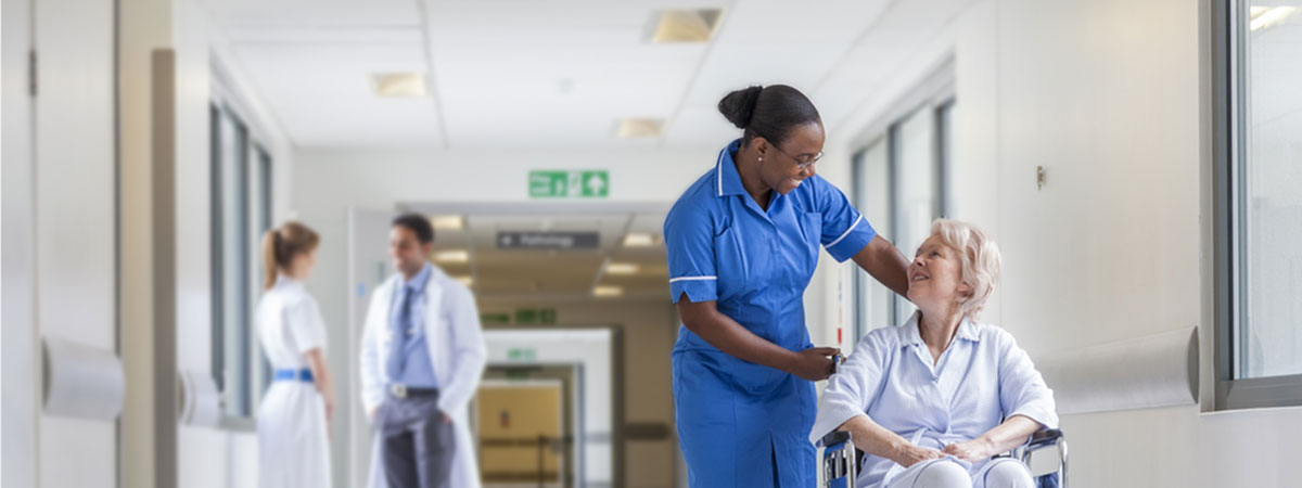 Healthcare worker in blue scrubs assisting an older patient seated in a wheelchair in a bright hospital corridor, with two other staff blurred in the background and signage overhead, clean clinical setting. Credit: Shutterstock